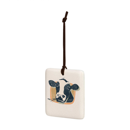 A square white tile hanging magnet ornament with a black and white cow on a tan background, displayed angled to the left.