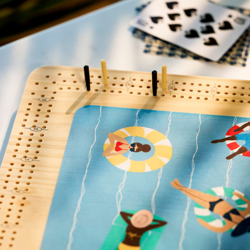 A wood cribbage game with a graphic image of people on floats in water. The game is sitting on a white table next to playing cards.