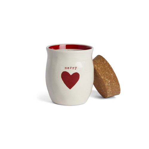 A cream ceramic container that says "merry" with a red heart and a cork lid. There is a poured candle inside, displayed with the lid off and leaning on the side.