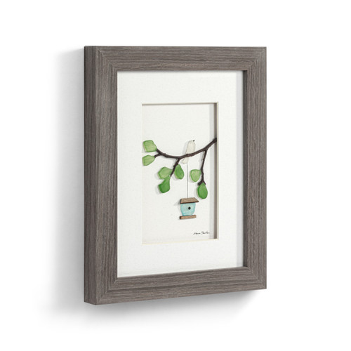 A gray wood framed image of a birdhouse hanging from a tree branch with a white bird perched on it made of pebbles, displayed angled to the right.