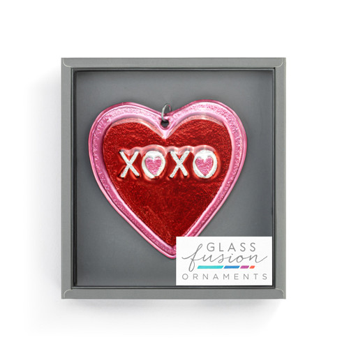 A red heart shaped glass ornament that says "XOXO" and is on a curved silver metal hanger, displayed in a packaging box.