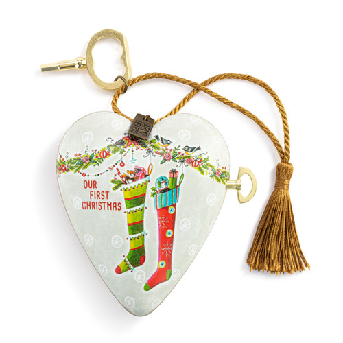 A white heart shaped musical sculpture with the saying "Our First Christmas" with an illustration of two full stockings. The heart has a gold tassel and gold key attached.