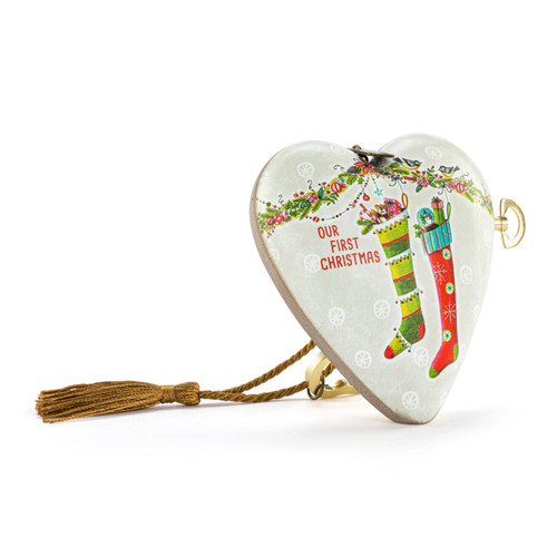 A white heart shaped musical sculpture with the saying "Our First Christmas" with an illustration of two full stockings. The heart has a gold tassel and gold key attached, displayed angled to the right.