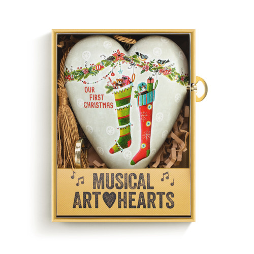 A white heart shaped musical sculpture with the saying "Our First Christmas" with an illustration of two full stockings. The heart has a gold tassel and gold key attached, displayed in a packaging box.