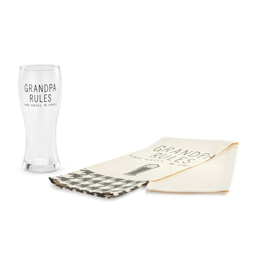 A clear pilsner glass that says "Grandpa Rules The Grill 'N Chill" next to a cream towel folded over itself with the same saying and a houndstooth pattern at the bottom.