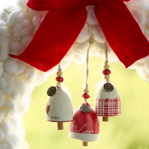 Three different red and white mini bells hung inside a white large knit wreath with a red bow hanging on a window.