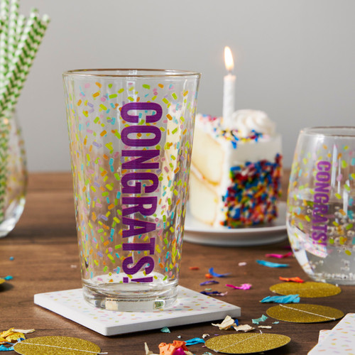 A celebration table with confetti, a slice of birthday cake and glass with confetti and the saying "Congrats!" on the outside.