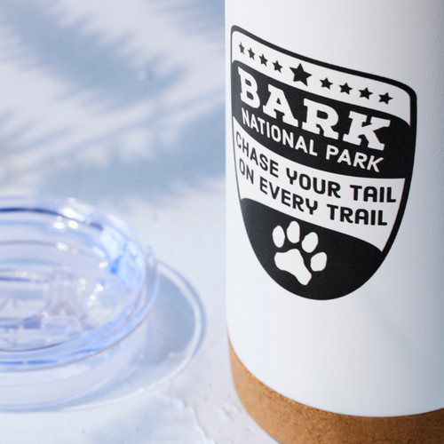 Close up view of a white tumbler with a cork base and the saying "Bark National Park Chase Your Tail on Every Trail" displayed on a light blue background.