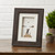 Brown grooved frame with white mat board and white background sits on wooden shelf. Pebbled person is dancing on dock with two small clouds overhead. White brick background