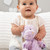 A close up image of a baby girl, sitting in front of two large pillows in a nursery scene, holding a soft, plush, purple and yellow T-Rex Rattle Blankie.