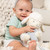 A close up image of a baby girl, sitting on a chunky knit ivory blanket in front of an ivory couch, holding a soft, plush lamb.