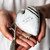 Close up of a woman holding a heart shaped sculpture in her hands that has a gold key and tassel attached. The heart says "Be Still & Simply Be" and features a white and black pattern inspired by ArtLifting artist Cheryl Kinderknecht.