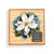 A square wood board for tic tac toe with a watercolor image of a white magnolia, displayed in a packaging box.