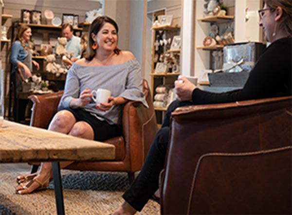 Two women sitting in a store drinking coffee with a store employee helping shoppers in the background