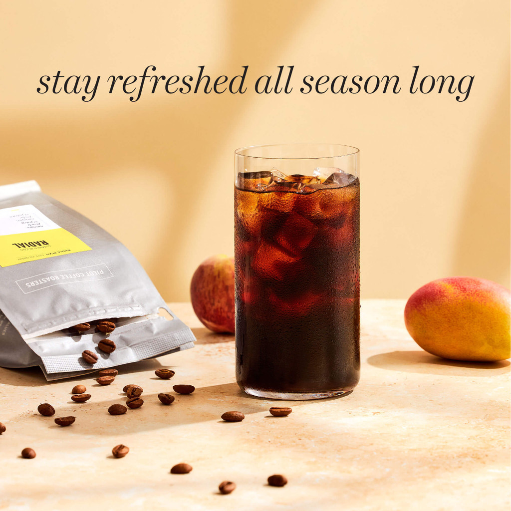 Text: Stay refreshed all season long. Image: cold glass filled with cold brew coffee and ice on a sandy stone countertop. A bag of radial sits on its side to the left with beans spilling out. A mango is sitting just to the right to emphasize the juicy mango notes that come through in this blend.
