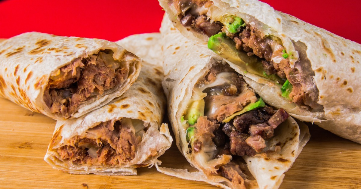 Cut burritos reveal beef, avocado, beans, and melted cheese as they rest on a wooden board against a red backdrop.