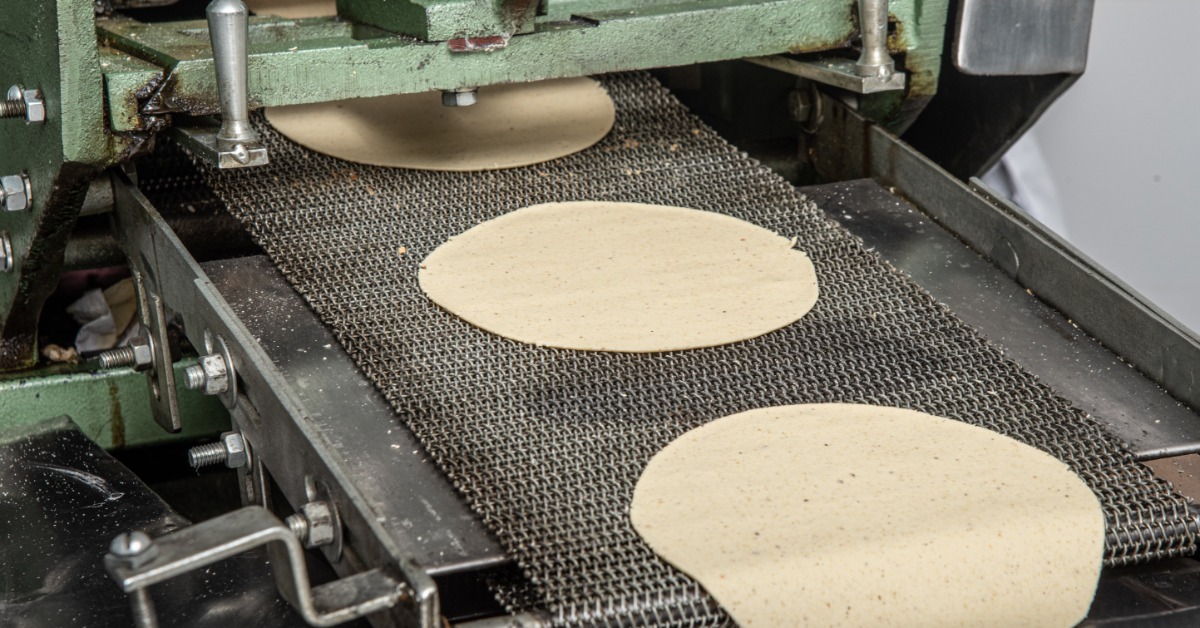 A commercial tortilla machine presses round tortillas onto a metal conveyor belt inside an commercial kitchen.