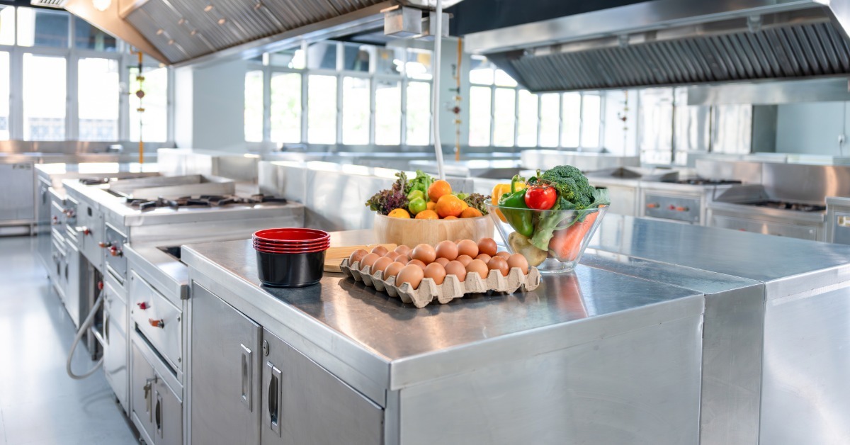 A commercial kitchen contains stainless steel cooking equipment with fresh vegetables and eggs on a central prep table.