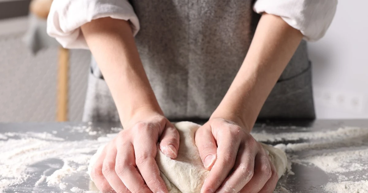 Mastering the Essential Art of Bread Proofing A baker presses a ball of bread dough with both hands on a flour-covered work surface in a bright kitchen.