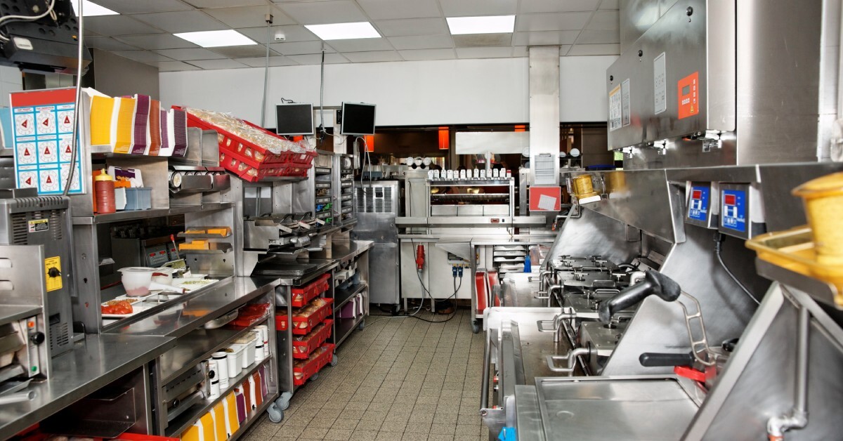 A bright commercial kitchen contains stainless steel counters, red crates, cooking stations, and organized shelving.