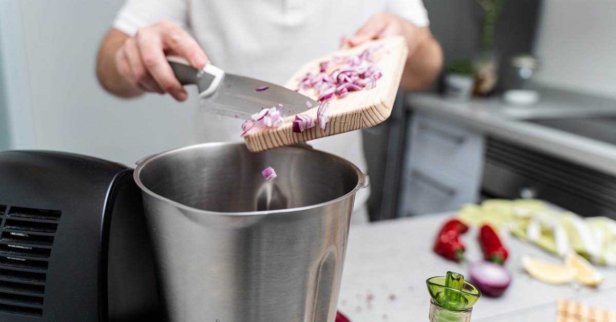 A chef adds chopped purple onion to a food processor. The onion sits on a wooden cutting board and they scrape with a knife.