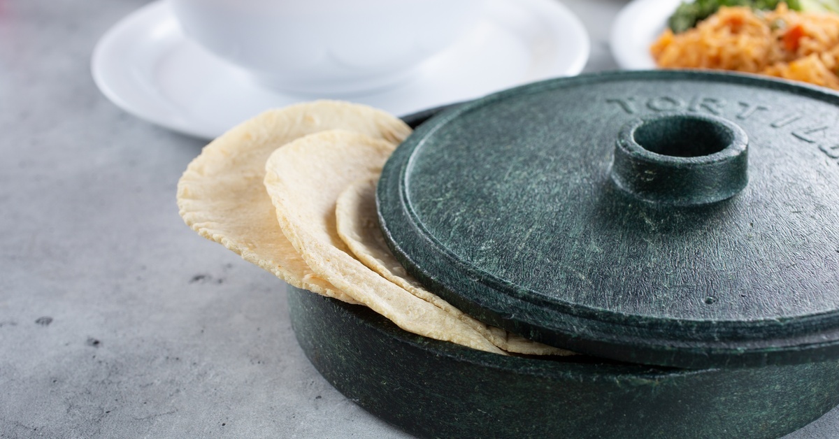 Some flour tortillas rest outside the edge of a small, dark green tortilla warmer. The warmer sits next to a plate of rice.