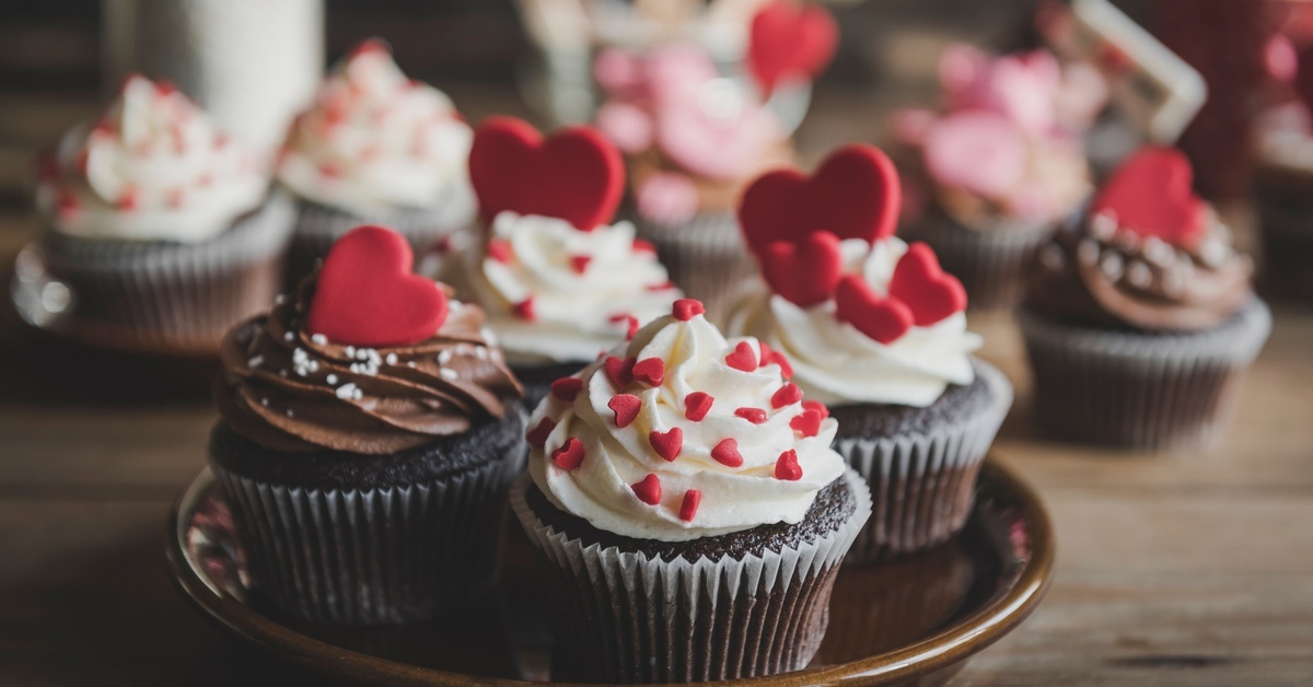 Chocolate cupcakes with white frosting and red heart sprinkles rest on a brown plate with more cupcakes in the background.