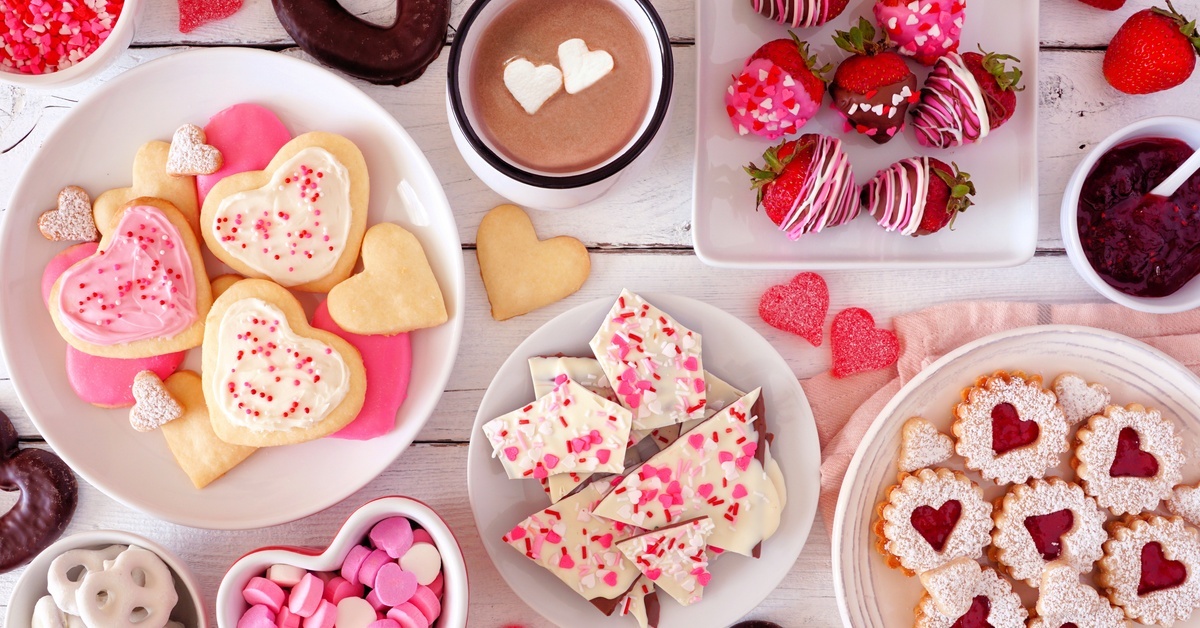 A top-down view of Valentine's treats, including heart cookies, chocolate-dipped strawberries, and candy sprinkles.