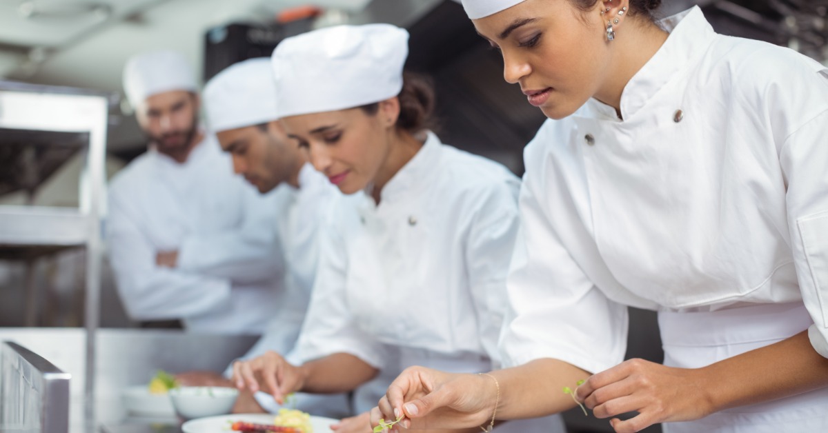 Best Practices for Portion Control in Restaurant Operations A group of chefs in white uniforms carefully plate dishes with garnishes at a stainless steel counter in a busy kitchen.