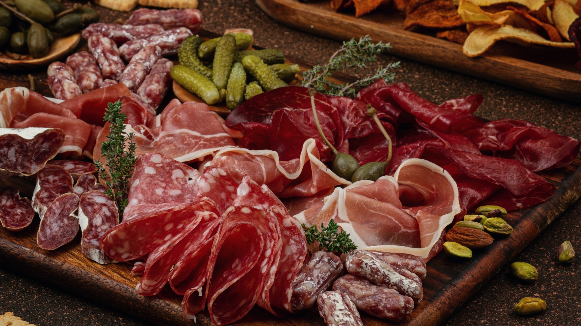 A wooden charcuterie board holds folded cured meats, pickles, crackers, chips, nuts, and herbs in a rustic spread.