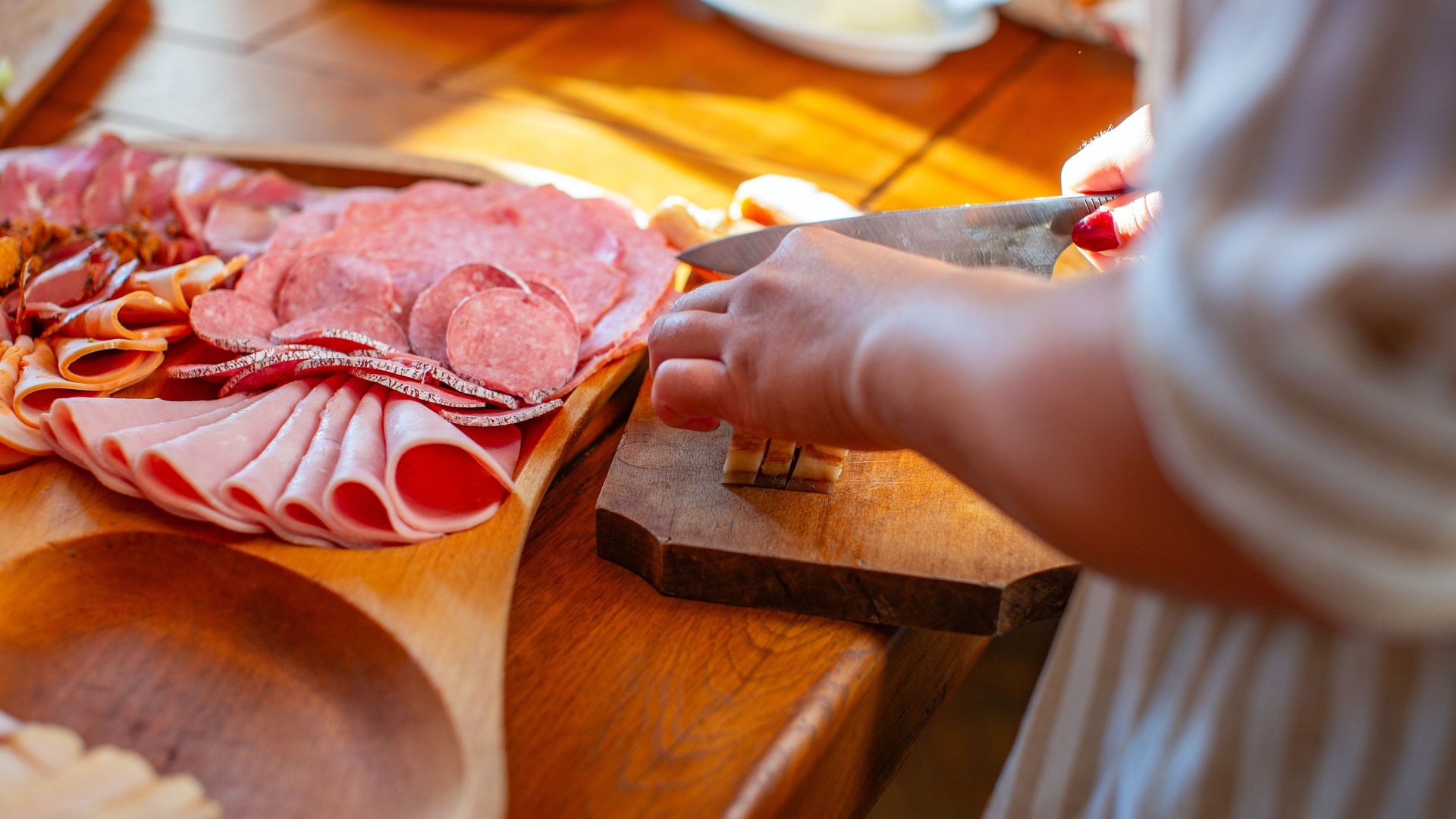 A cook slices cheese beside a wooden board filled with folded deli meats and salami on a sunlit table.