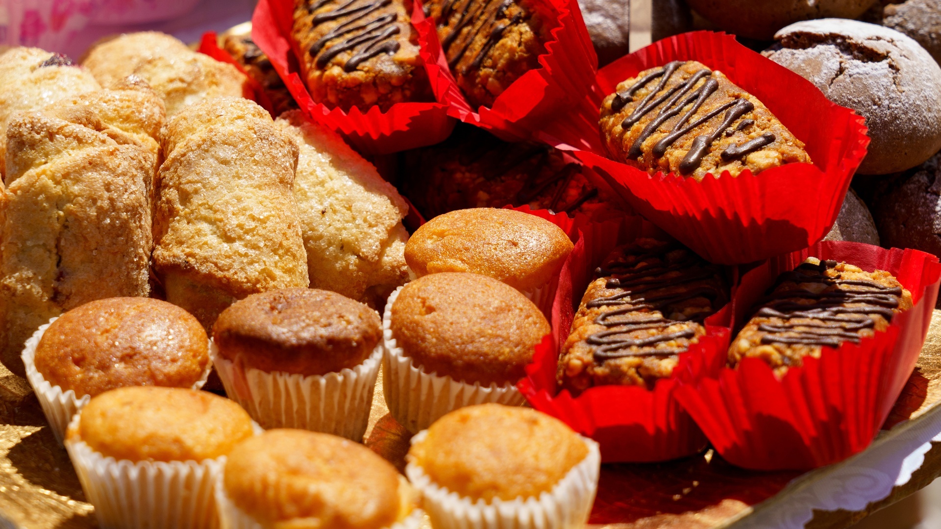 A platter holds golden muffins, sugar-dusted pastries, and chocolate-drizzled baked goods in red paper liners.