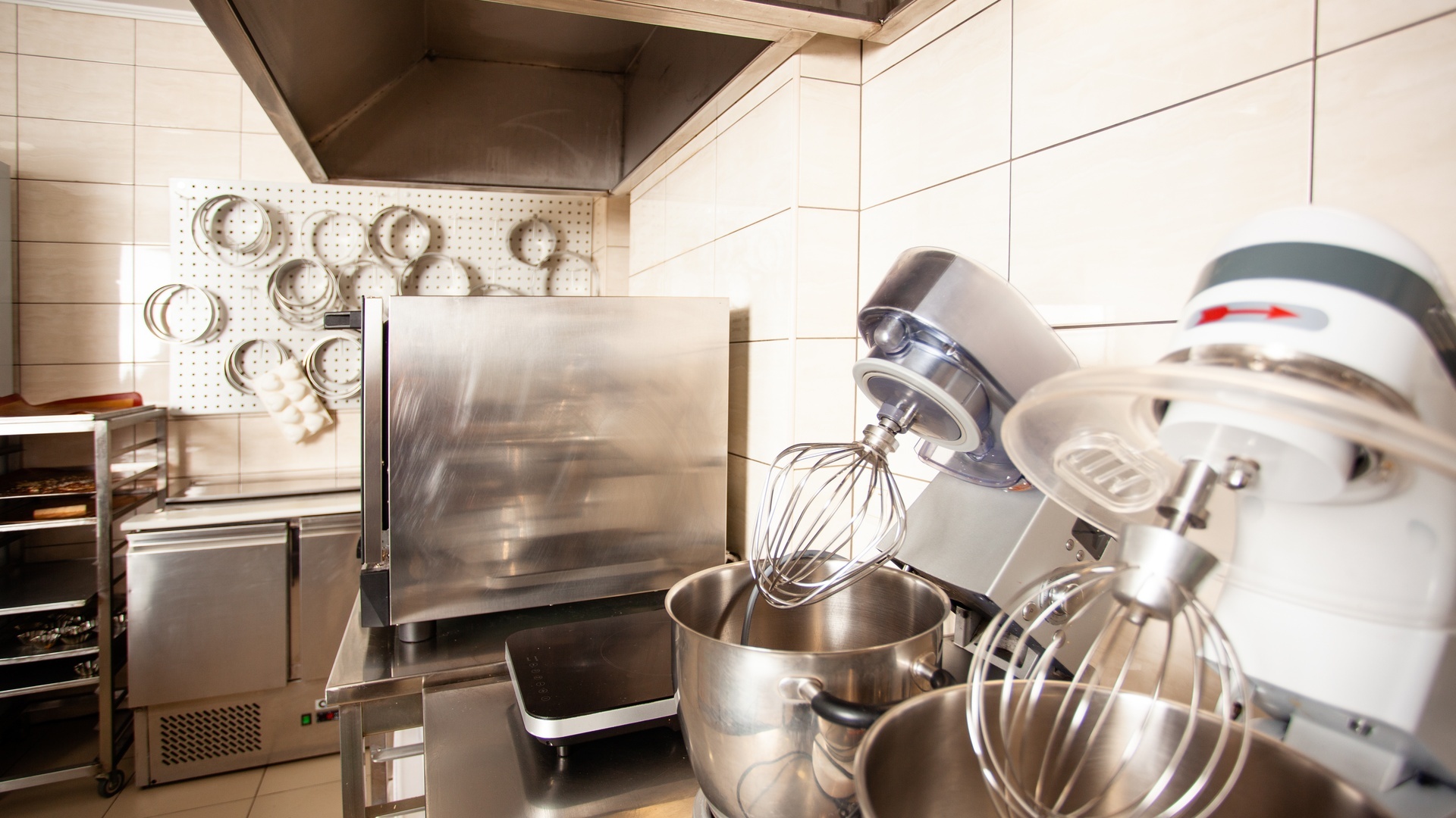 Two stand mixers hold whisk attachments beside a stainless oven and hanging pans in a bakery kitchen.