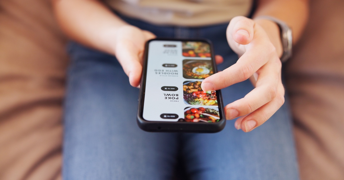 A woman holds a smartphone showing a ghost kitchen's menu, with her finger scrolling through colorful food options.