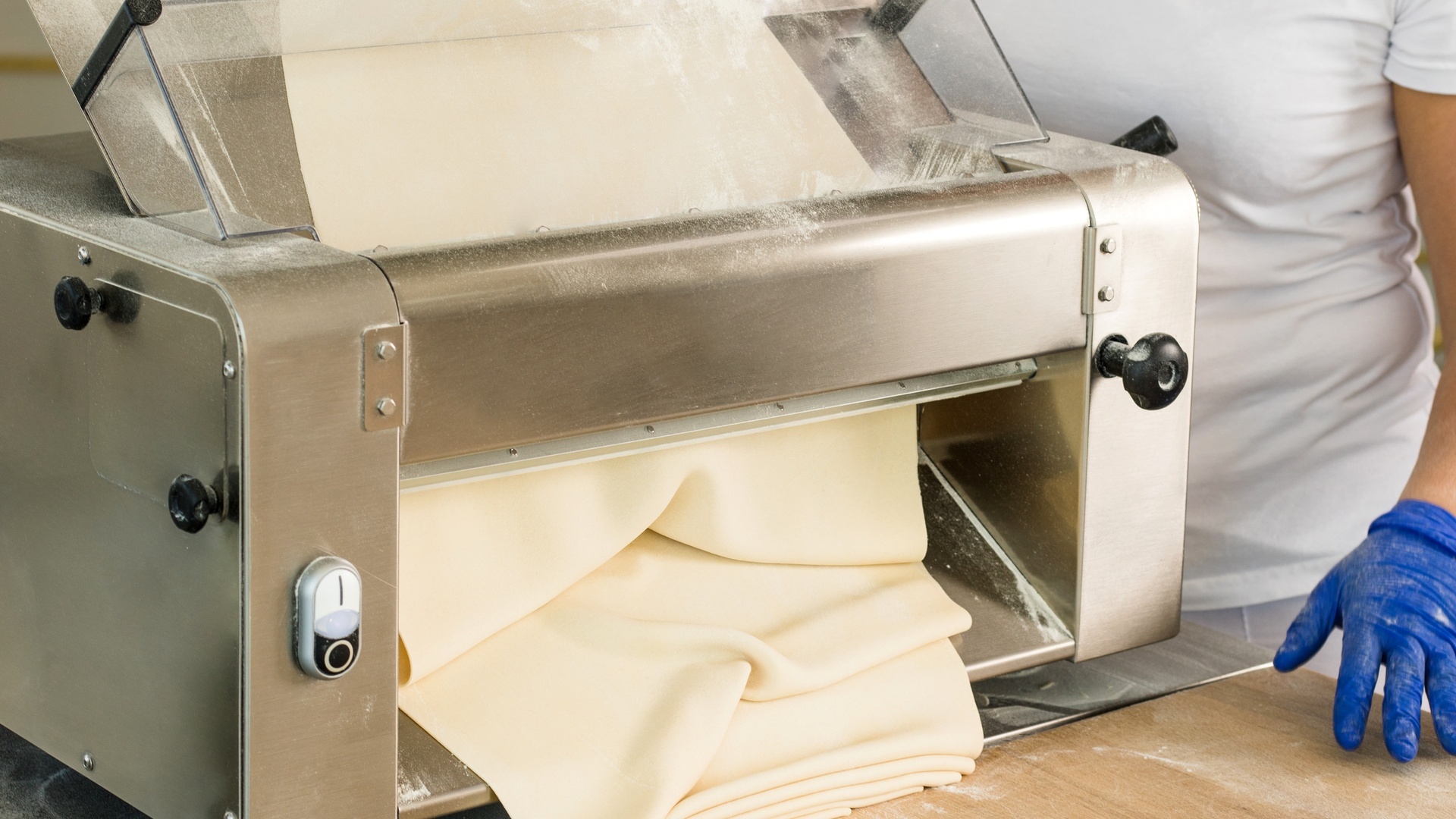 A baker feeds dough through a commercial dough sheeter with gloved hands on a floured table, creating thin folded sheets.