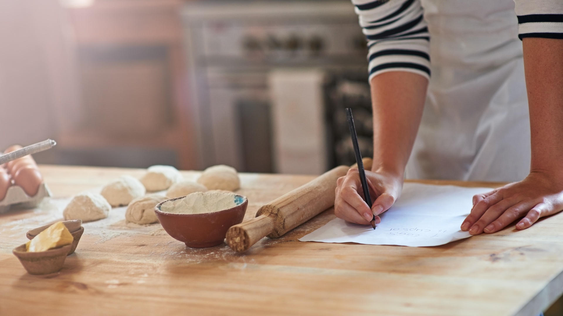 A baker writes down recipes at a baking table with dough balls, a rolling pin, and a bowl of flour nearby.