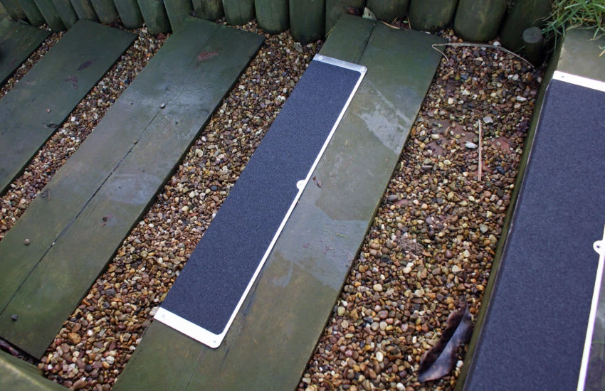 Heskins L-Shape Bolt Down Anti-Slip Plate close-up on wet timber decking — pre-drilled countersunk mounting holes accept #10 or #12 stainless screws or Tapcon-style concrete anchors