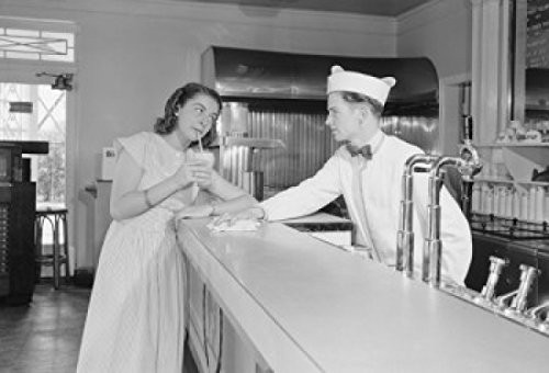 Young woman drinking milkshake at bar counter with barman Poster Print (18 x 24)