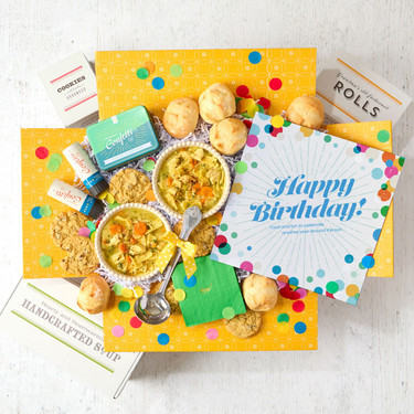 Overhead view of a birthday-themed gift arrangement with two bowls of soup, rolls, cookies, a container labeled “Handcrafted Soup,” a metal ladle with a yellow ribbon, green birthday napkins, confetti poppers, colorful confetti, and a card that reads “Happy Birthday!”