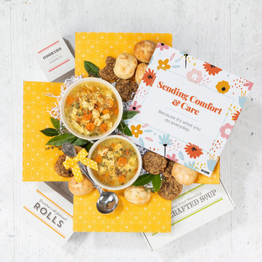 Overhead view of a yellow gift box arrangement with two bowls of soup, rolls, cookies, a container labeled “Handcrafted Soup,” a metal ladle with a yellow ribbon, and a floral card that reads “Sending Comfort & Care.”