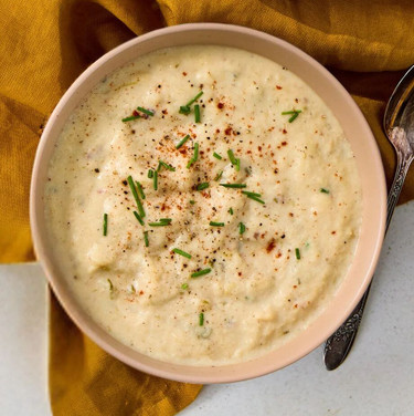 Close-up of a bowl of loaded baked potato soup topped with herbs and seasoning, set beside a spoon and a yellow cloth.