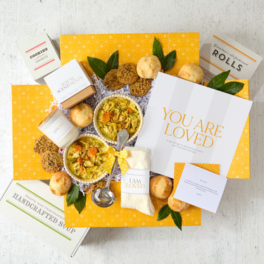 Overhead view of a yellow gift box arrangement with two bowls of soup, rolls, cookies, a container labeled “Handcrafted Soup,” a metal ladle, a candle, a pair of cozy socks, and a card that reads “You Are Loved.”