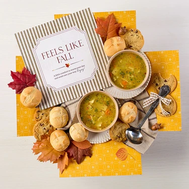 Overhead view of a fall-themed arrangement with two bowls of soup, rolls, cookies, a box labeled “Handcrafted Soup,” a box of rolls, a metal ladle with a yellow ribbon, autumn leaves, and a card that reads “Feels Like Fall.”