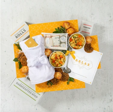 Overhead view of a relaxation-themed gift arrangement with two bowls of soup, rolls, cookies, a box labeled “Handcrafted Soup,” a robe, self-care items in a toiletry bag, a metal ladle with a yellow ribbon, and a card that reads “Relax.”