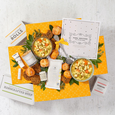 Overhead view of a yellow gift box arrangement with two bowls of soup, rolls, cookies, a container labeled “Handcrafted Soup,” a metal ladle with a yellow ribbon, a candle, small self-care items, and a card.