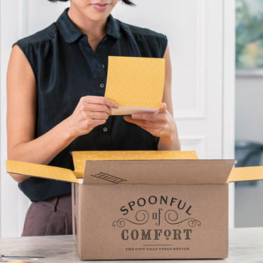 Person opening a Spoonful of Comfort box and holding a yellow card, standing at a table indoors.