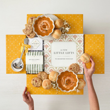 Top-down view of an open gift box with bright yellow patterned flaps containing bowls of soup, bread rolls, cookies, and a honey dipper. Two hands reach in to pick up the rolls, with a striped tea towel and a card in the center that reads “A Few Little Lifts.”