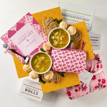 Overhead view of a comfort food gift package with two bowls of soup, dinner rolls, cookies, and coordinating pink and yellow packaging.