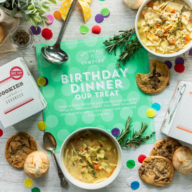 Overhead view of a birthday-themed food gift arrangement with bowls of soup, cookies, rolls, and packaged cookies surrounding a green card that reads “Birthday Dinner Our Treat,” with colorful confetti scattered on the table.