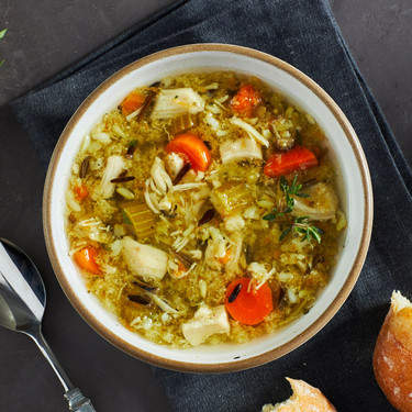 Close-up of a bowl of chicken and wild rice  soup with carrots, herbs, and shredded chicken, placed beside a spoon and a piece of bread.